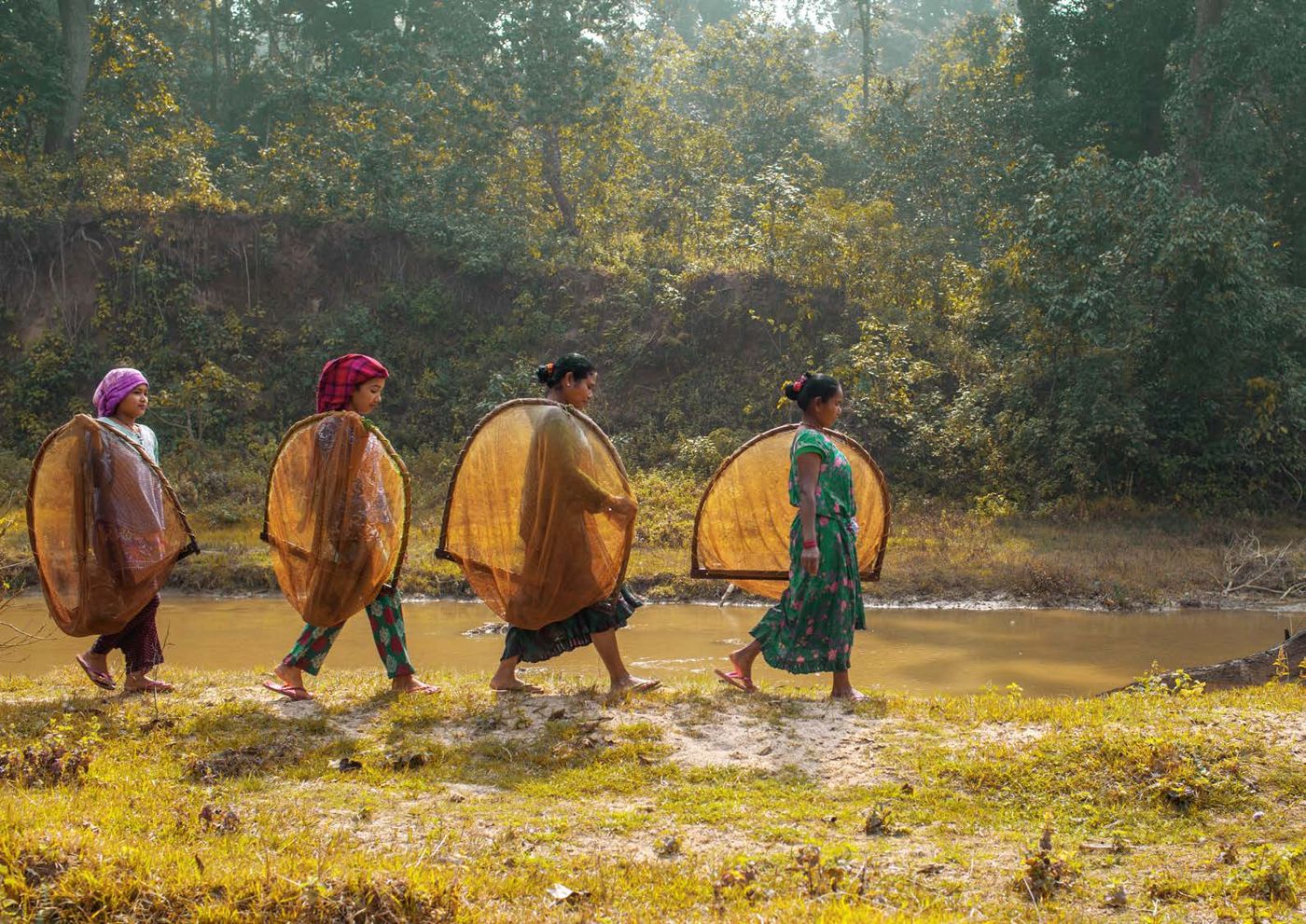 Four women walk along a riverbank carrying large round fishing nets.